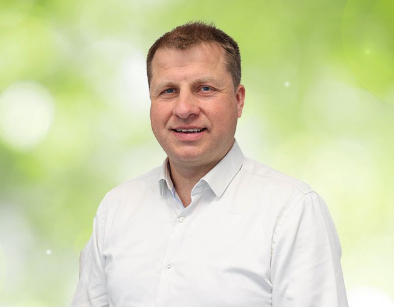 Ein lächelnder Mann in einem weißen Hemd vor einem unscharfen, hellgrünen Hintergrund.